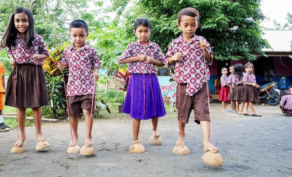 Four children in matching button-up shirts with brightly colored geoemetric patterns are playing a game where they walk on halved coconut shells, which are attached to ropes that they are holding with their hands.