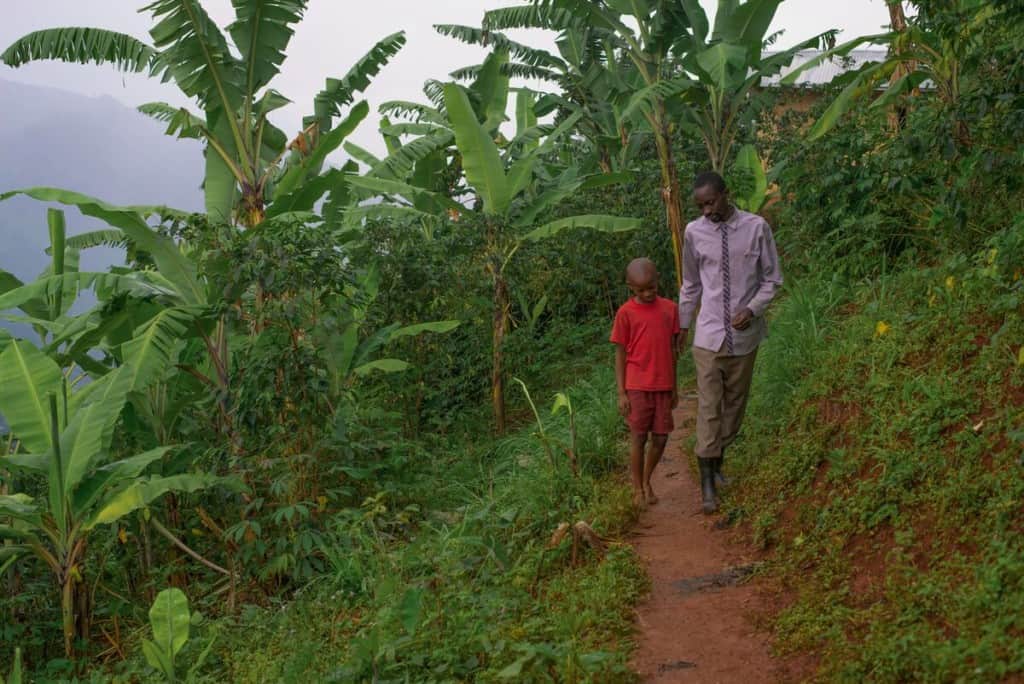 A father and son in Uganda walk down a narrow, red dirt trail leading through thick green foliage. The boy is wearing a red shirt and red shorts. The father is wearing a purple shirt and rainboots.