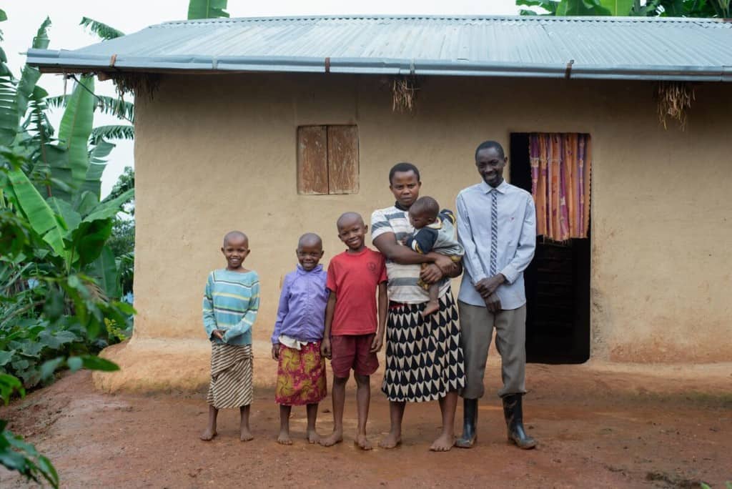 A family of six people stands outside their home in Uganda. The mother is holding a baby. The father is wearing rain boots. The mother and children are wearing colorful clothing and are barefoot.