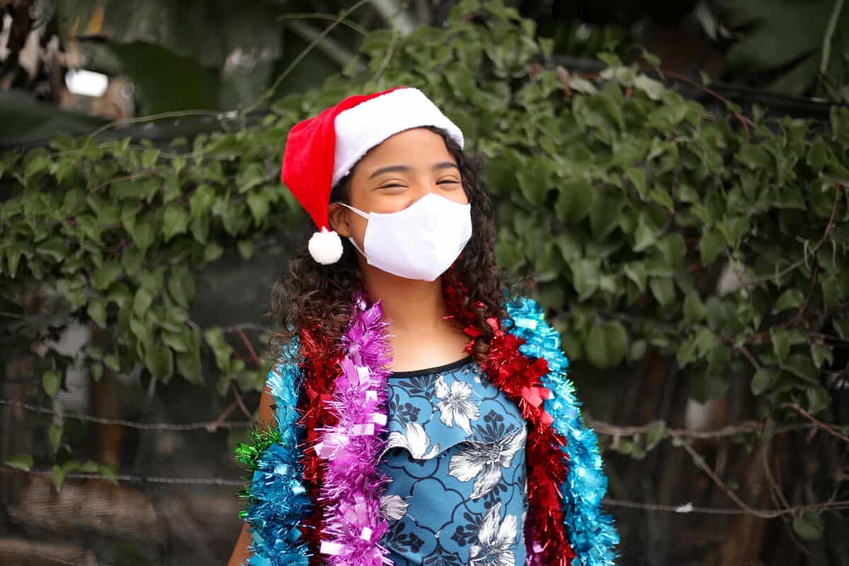 A girl in Brazil wears a white face mask, Santa hat and sparkly garlands around her shoulders.