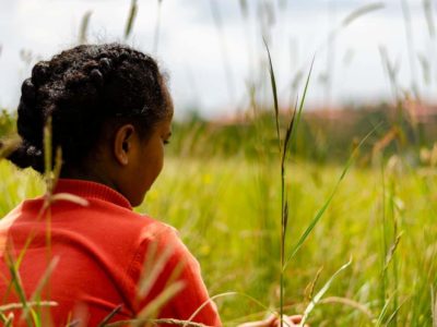 A girl wearing braids and a red shirt sits in a grassy field