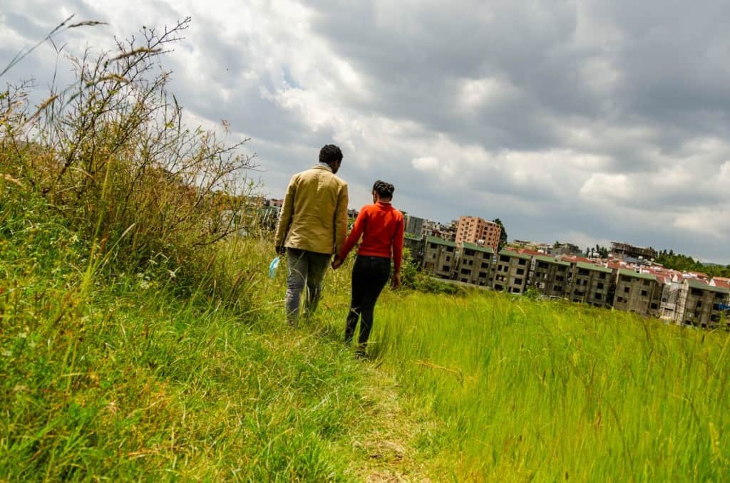 A man and a girl walk through a grassy field holding hands. There are rows of buildings in the background.