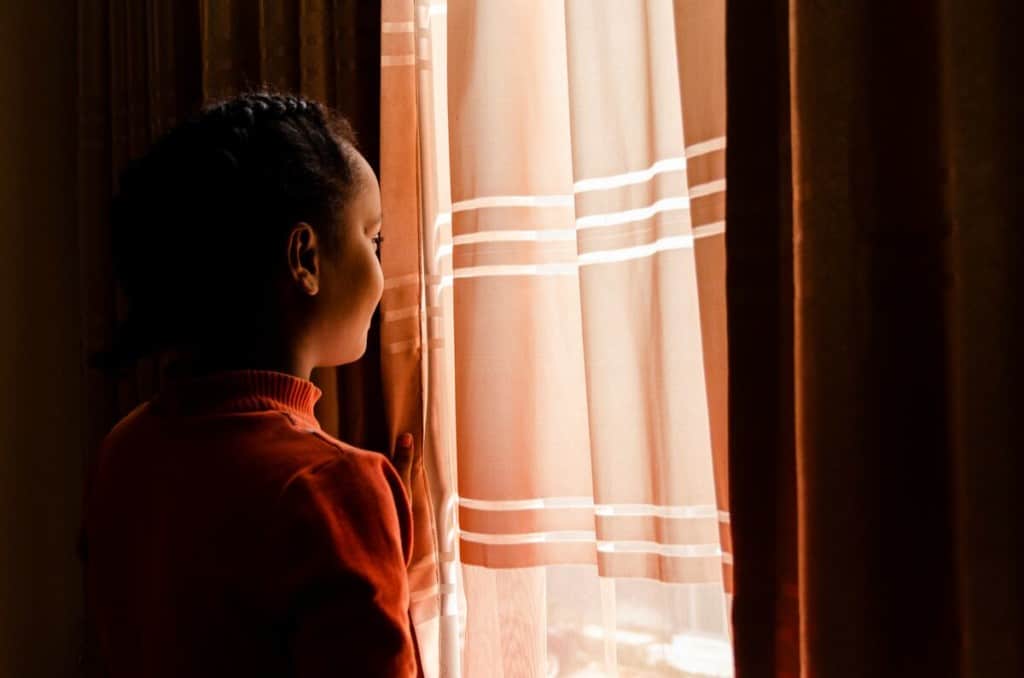 A girl looks out a window that is covered by pink-orange curtains.