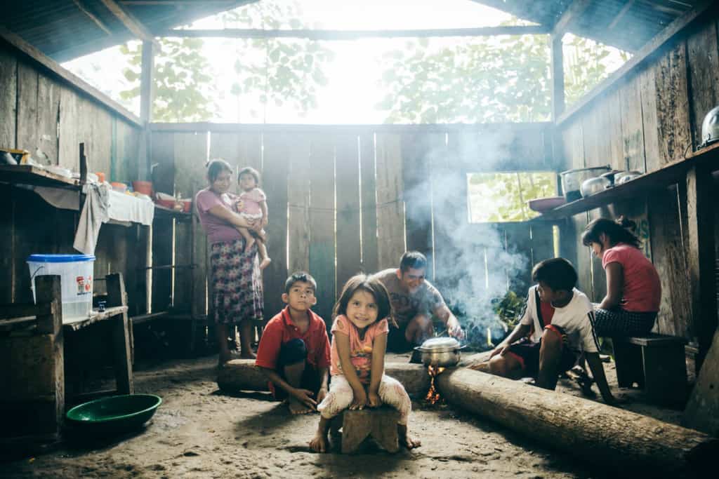 A family of boys, girls, mother holding a baby and father are inside a house with wood plank walls. A man tends to a silver pot cooking over a flame between three logs and a smiling girl in a pink shirt and white pants sits on a stool. Smoke rises from the fire.