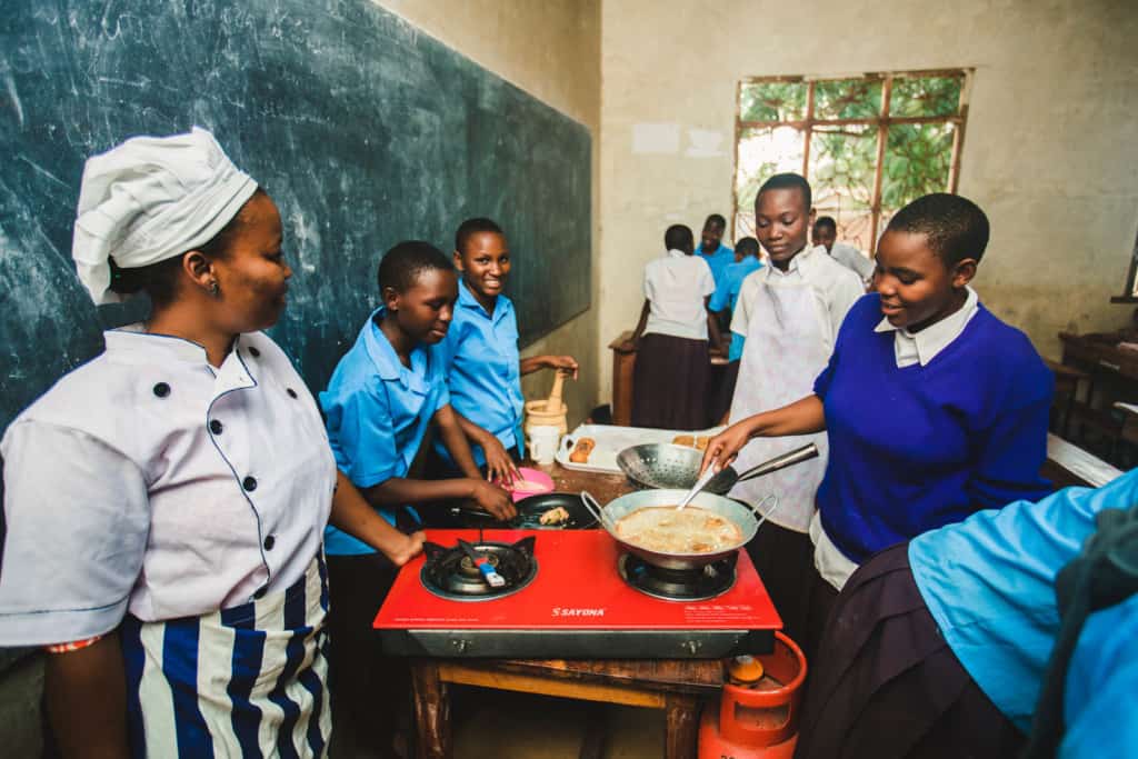 A woman in a blue and white striped apron, white chef hat and jacket stands next to a red cooktop resting on a table. A student in a blue sweater stirs a pan of food. There is a propane tank on the floor and a chalkboard along the wall. Students in blue uniform shirts stand in the background. 