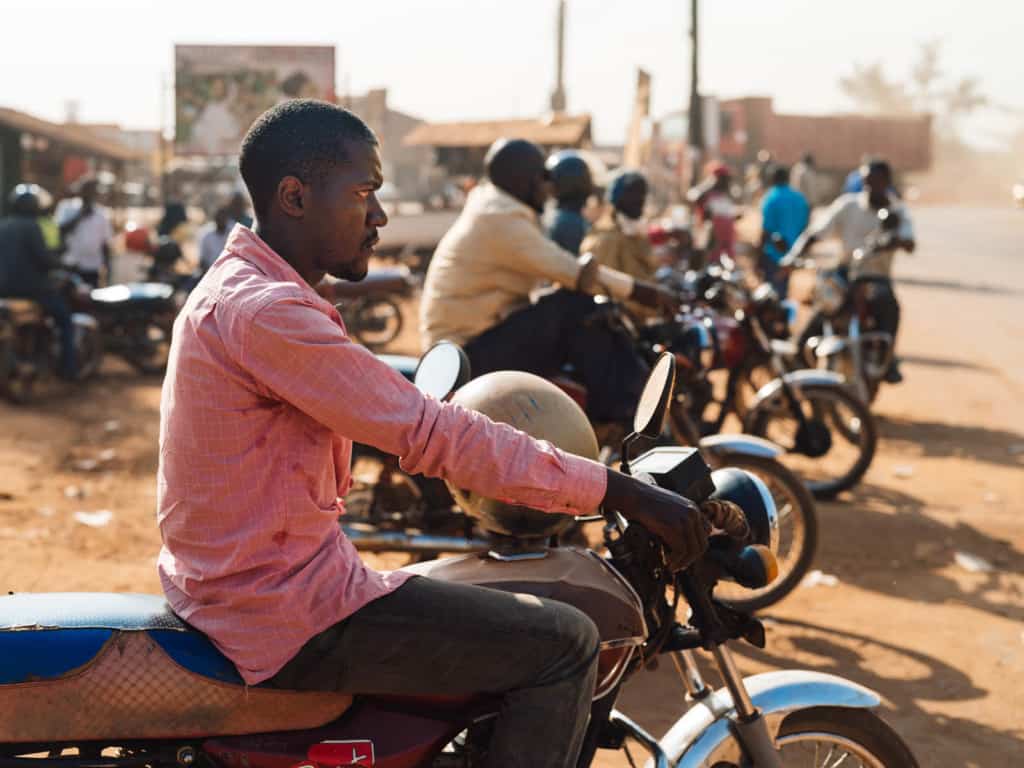 Boda driver (motorcycle taxi) is wearing a pink shirt and sitting on a motorcycle. There are more men on motorcycles in the background. Driving a boda is their primary source of income.