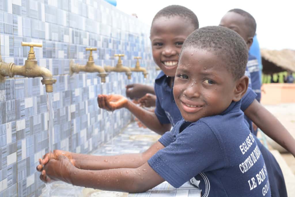 Girl Mary is wearing her Compassion uniform, a blue shirt and skirt. She is getting water from a river with a green bowl and is pouring it into a larger blue container.