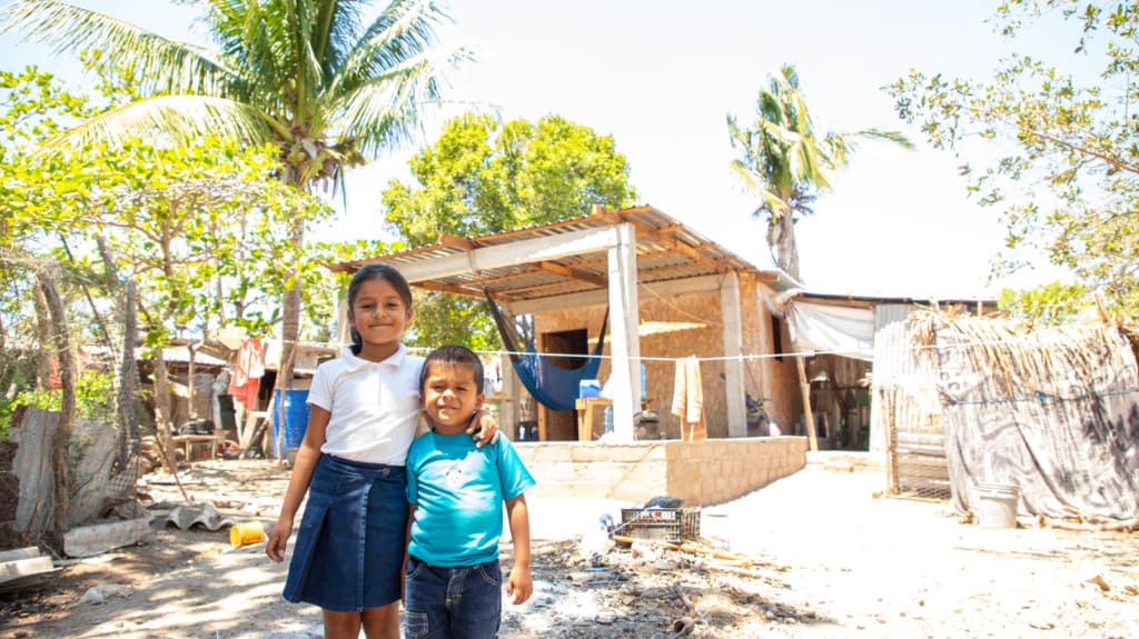 A girl wearing a blue skirt and a white shirt. She has her arm around her brother who is wearing a blue shirt. They are standing outside their new home.