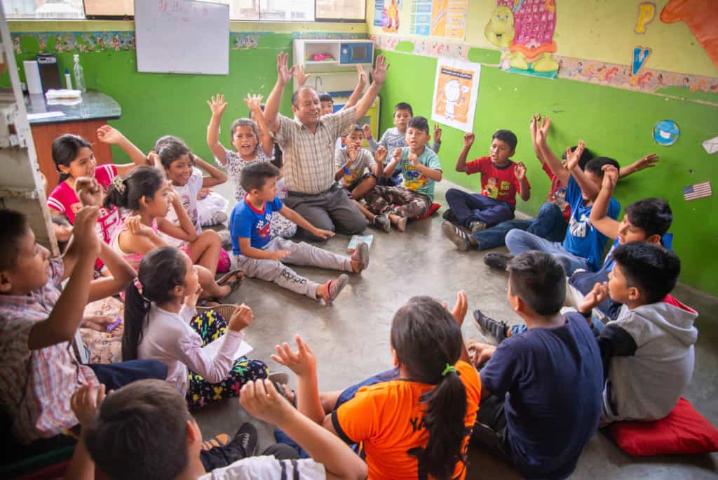 Pastor Agapito is praying with the children in one of the church classrooms. The walls are green with colorful decorations. Pastor Agapito and the children are raising their hands in the air.
