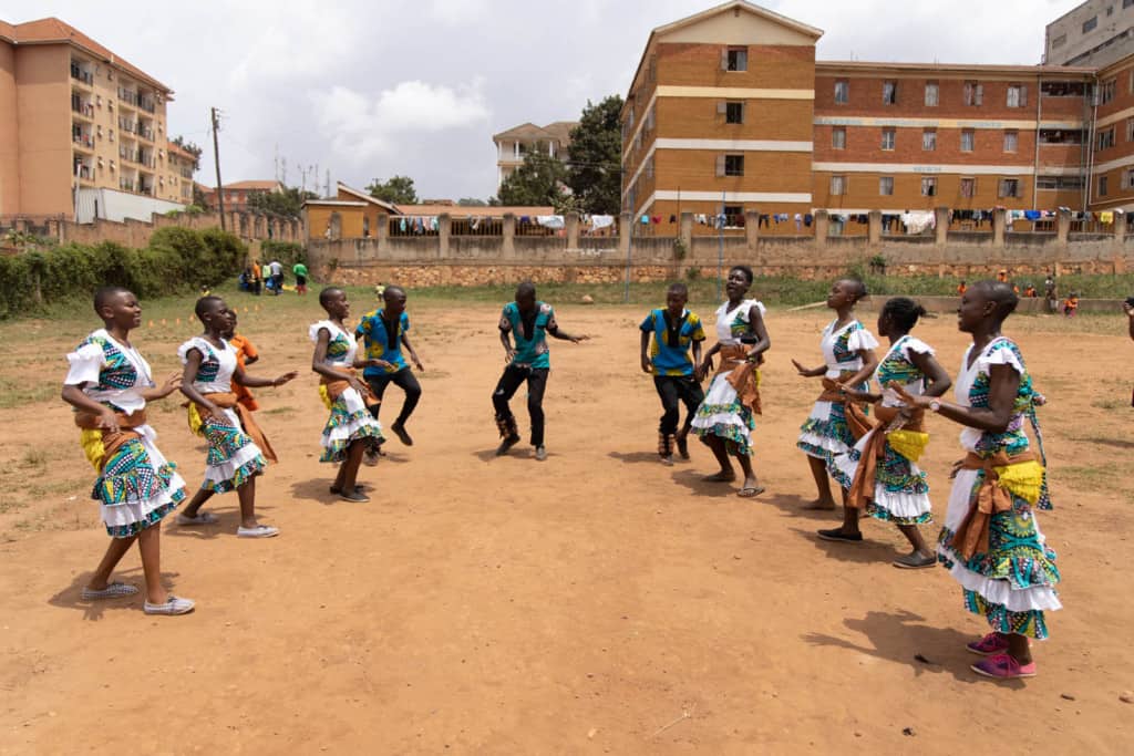 Group of children dancing in a dirt field in a city.