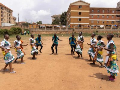 Group of children dancing in a dirt field in a city.