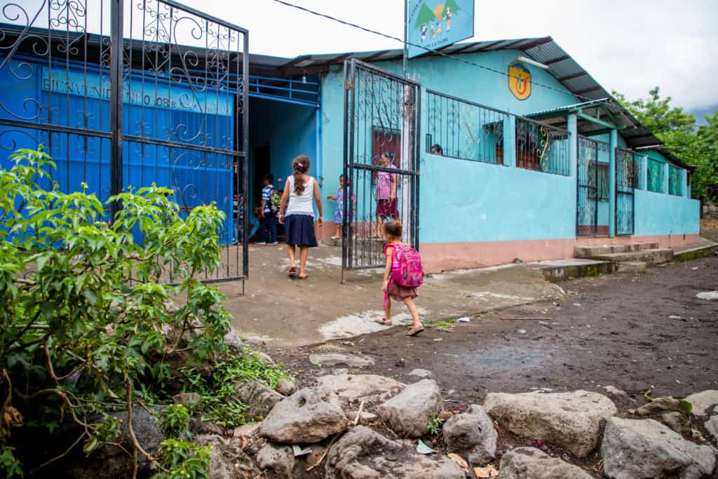 Girl walking into the Compassion project carrying a pink backpack. The building is blue and there is a wrought iron fence surrounding it.