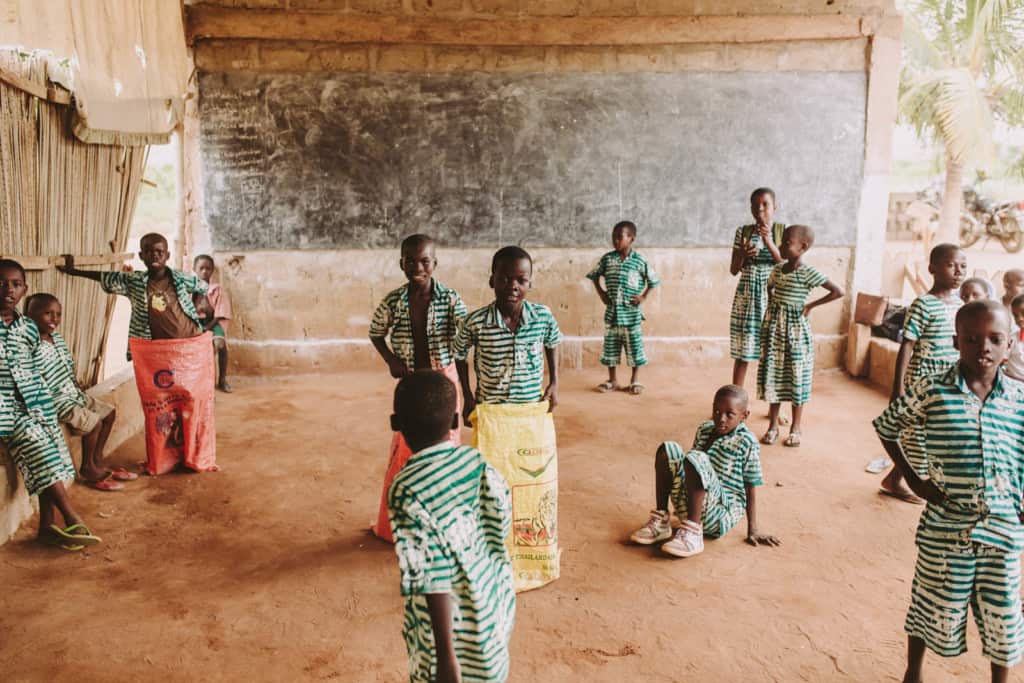 A group of children boys and girls students wearing school center uniform green and white dresses shirts and shorts stand inside a covered open room at the center playing games fun activities participating in games play and activity time with some of the children in large yellow or red sacks bags games. 