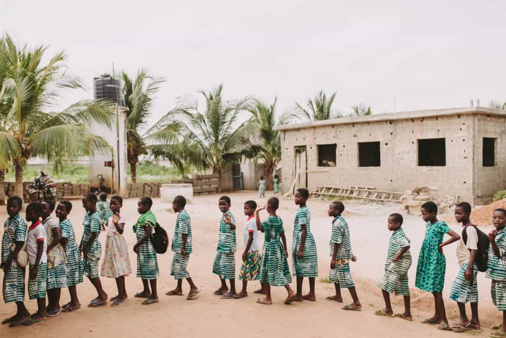 A group of children students boys and girls some wearing school center uniform green and white striped or polka dot dress shirts and pants stand in a single file line outside the center in the yard. A few are looking and waving with their hands. 