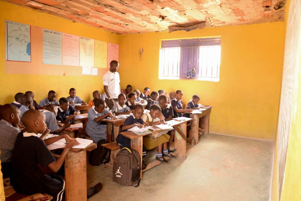 A classroom full of children. The teacher is standing among the desks. The walls are yellow.