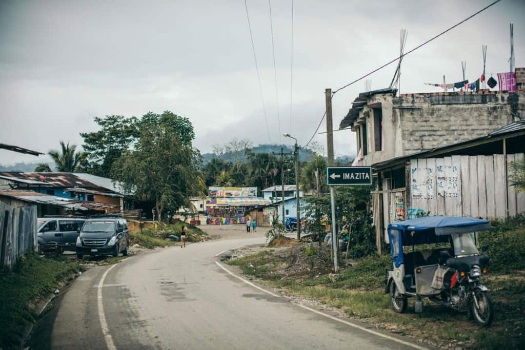 Cars and a blue three wheel motorcycle cab border a road. There are buildings and people walking in the street in the background.