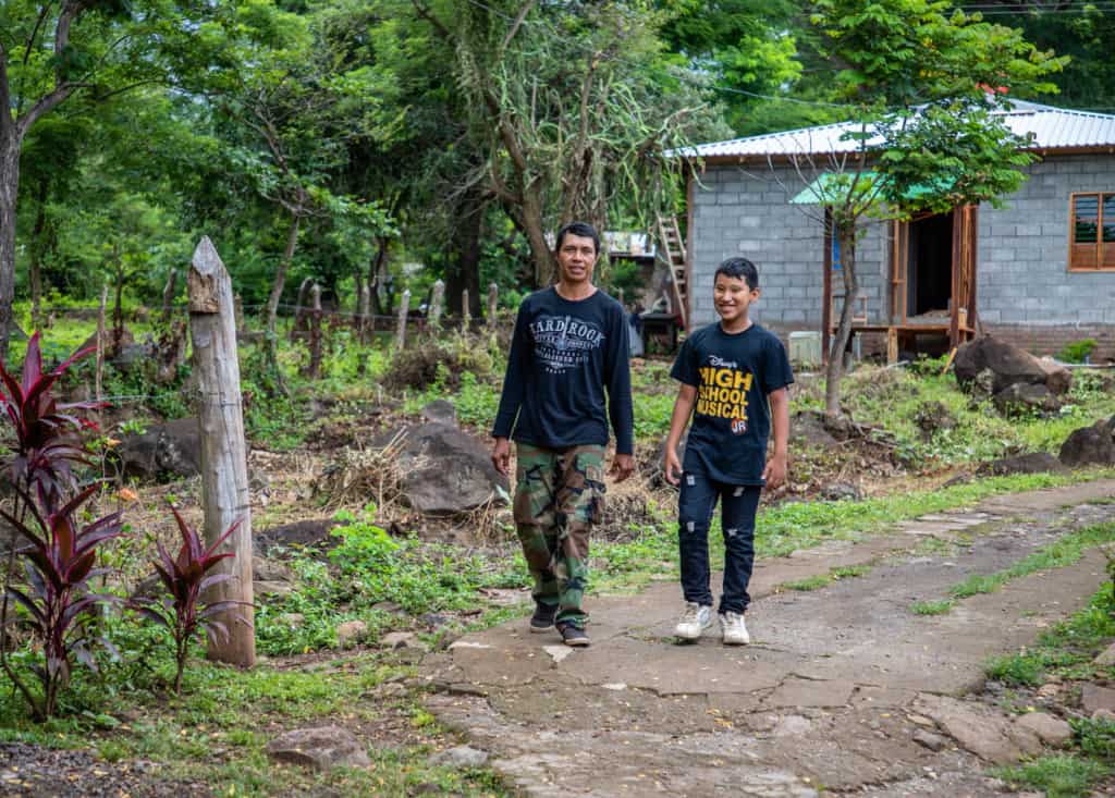 Boy wearing a black shirt, ripped jeans, and white tennis shoes. His father (left) is wearing a black, long sleeved shirt, camo pants and black shoes. They are walking together down a dirt road with their house behind him.