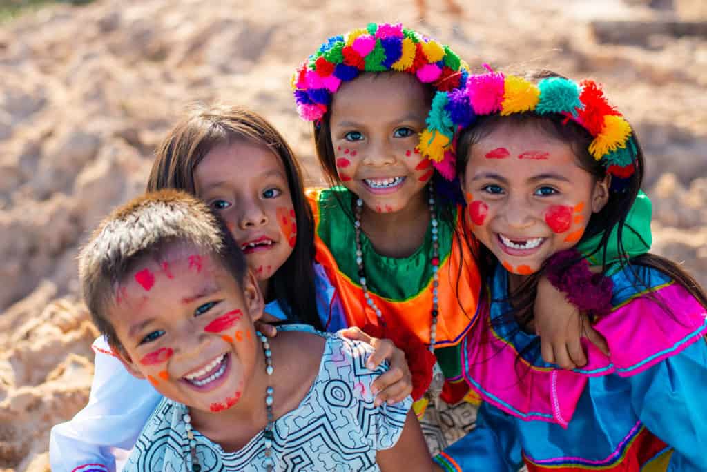 Four children are dressed in colorful outfits and have red paint on their faces. They are standing outside with their arms around each other.