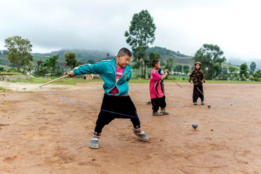 A boy wearing a blue jacket and black pants hurls a spinning top on the dirt ground outside.