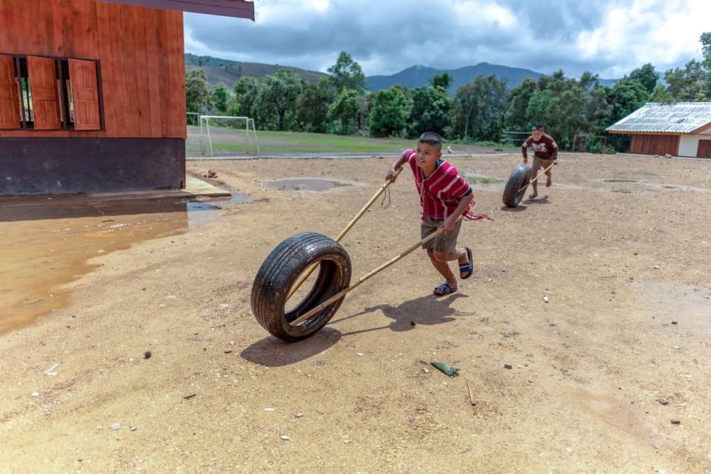 A boy wearing a red and white shirt, shorts and sandals uses bamboo poles to hold up the wheel of a car tire while running outside. Another boy is running after him with his own tire and bamboo poles.