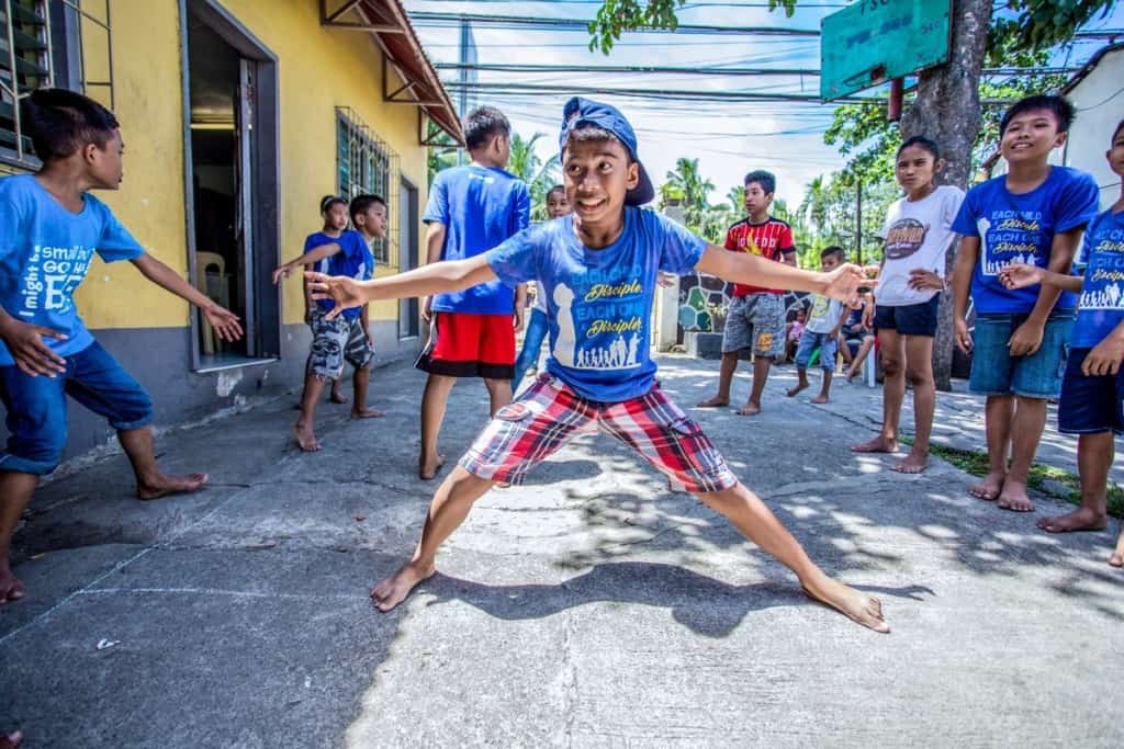 A group of children in Compassion's program is playing the game patinero, with one boy straddling two boxes drawn with chalk and holding his hands out in a T. Most children are wearing blue T-shirts and are barefoot.