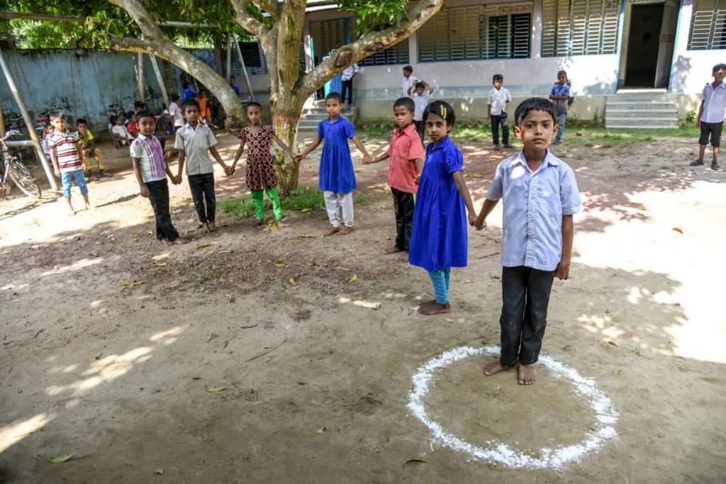 Seven children stand in a line outdoors in Bangladesh. They are holding hands. One boy stands in a circle drawn from white chalk. The children are playing the game golla choot