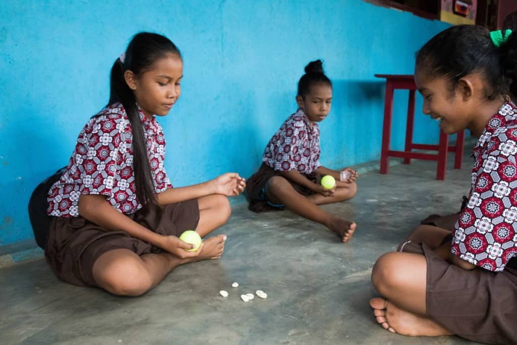 Three girls sit on a clean concrete floor in front of a blue wall. They are playing the game of bola bekel. Two are holding yellow balls in their hands. There are six small seashells on the floor between them.