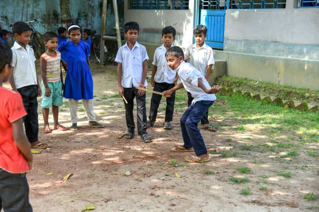 A group of young children in the Philippines stands in a semi-circle watching a boy swinging a stick at a smaller stick, playing the game chenti-benti