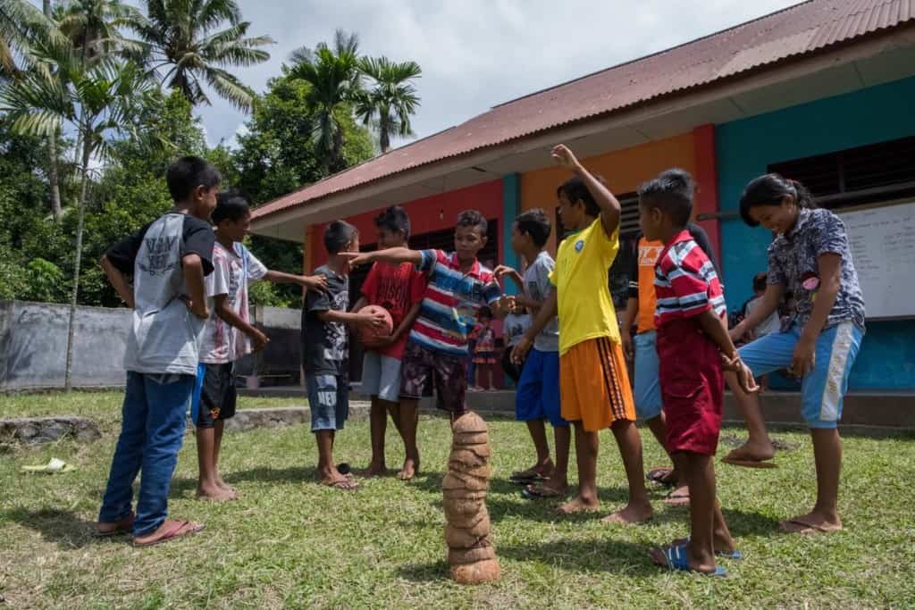 A group of children in Thailand is playing a game. They're wearing colorful clothing. One is holding a ball. A stack of coconut half-shells is seen on the grass in front of them. A colorful building is behind them.