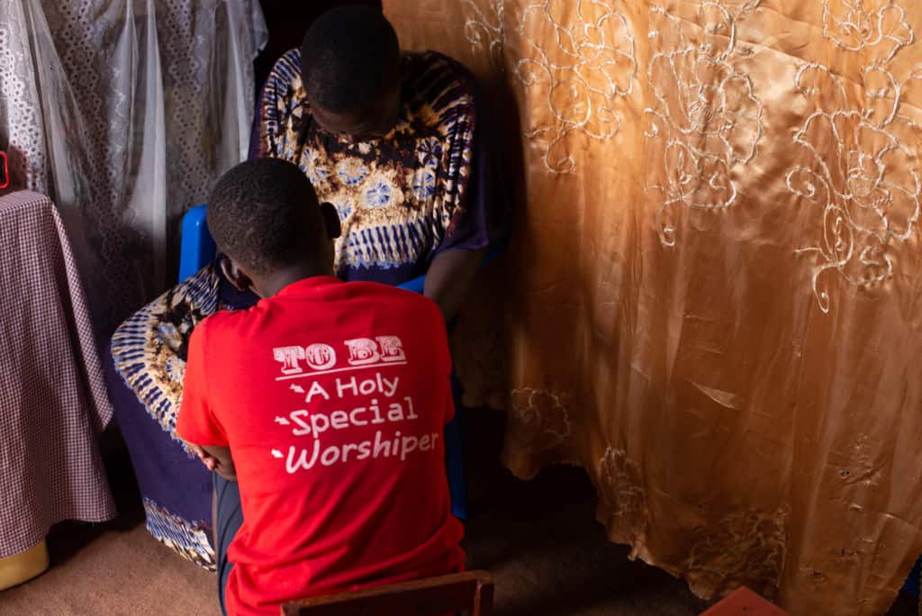 Rita is wearing a red shirt. She is sitting on the ground and is praying with her mother, Margaret, who is sitting behind her in a blue plastic chair.