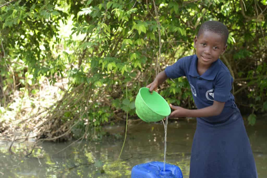 Girl wearing her Compassion uniform, a blue shirt and skirt. She is getting water from a river with a green bowl and is pouring it into a larger blue container.