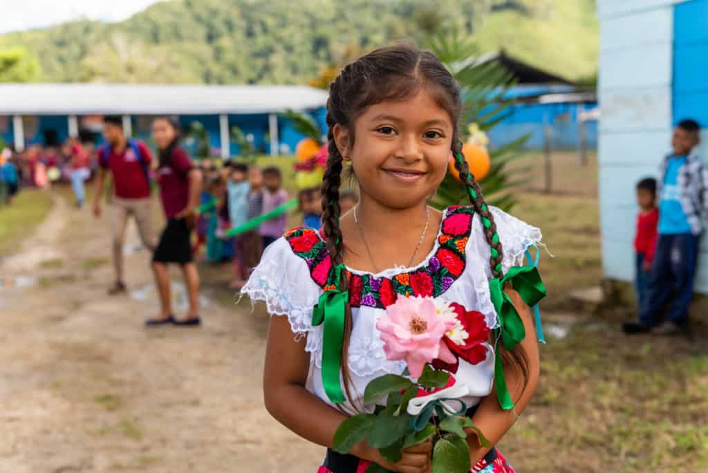 A girl wearing a white shirt is holding a bouquet of flowers. She is standing outside. There are other people in the background.