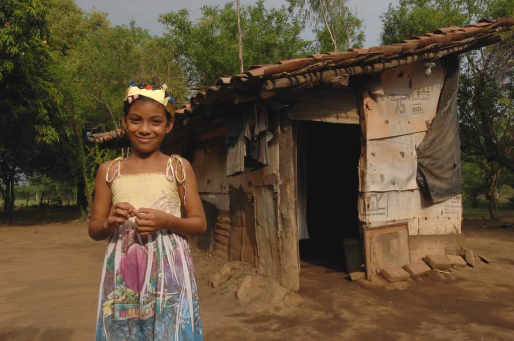 Girl standing in front of home that is constructed with cardboard pieces and black tarp. The roof has tile pieces. She is wearing a sun dress and a gold crown on her head.