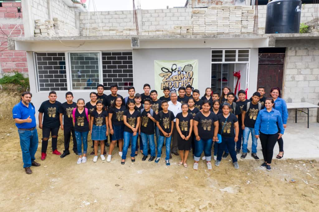 Youths (in black shirts) and tutors (in blue shirts) involved in the backpack production project are standing in front of the new center that was built for this project. The building is white.