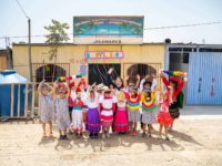 Compassion Peru's national director posting with children in front a church. The children are smiling and waving while wearing brightly colored clothing.