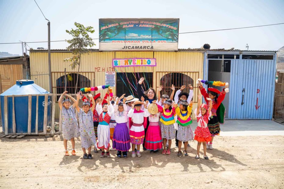 Compassion Peru's national director posting with children in front a church. The children are smiling and waving while wearing brightly colored clothing.