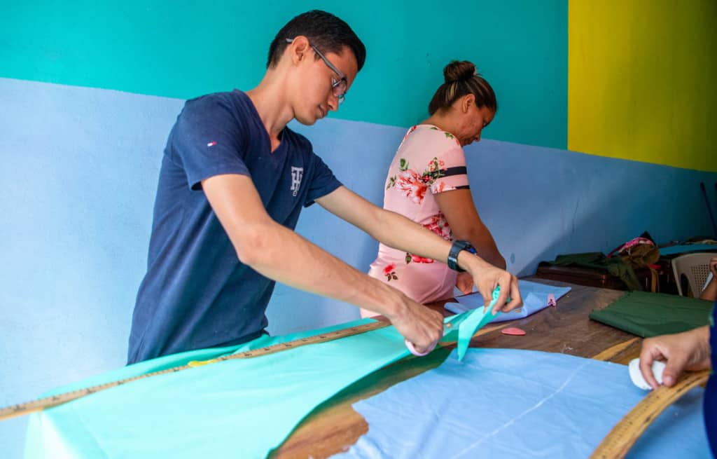 Victor, in a blue shirt, is standing at a work table cutting a piece of green fabric.