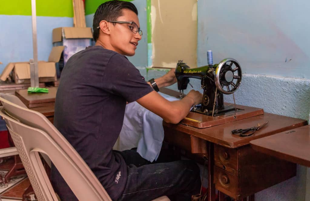 Victor, in a black shirt and jeans, is sitting at a sewing machine table in the classroom used for the sewing workshop. The walls are green and blue.