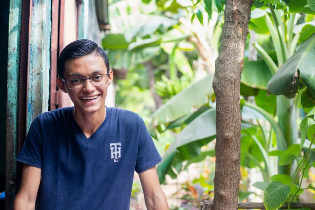 Victor, in a blue shirt, is smiling and standing outside his home. There are tropical trees next to him.