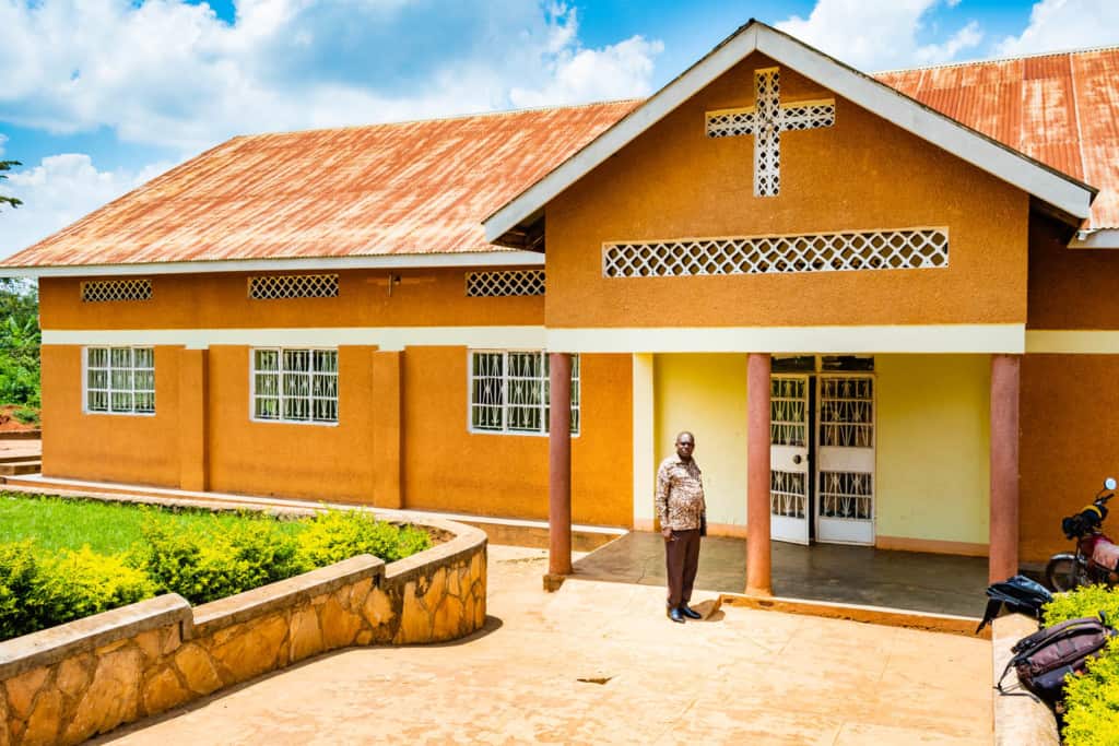 Pastor Cyrus is wearing a brown patterned shirt and brown pants. He is standing in front of a mustard yellow church.