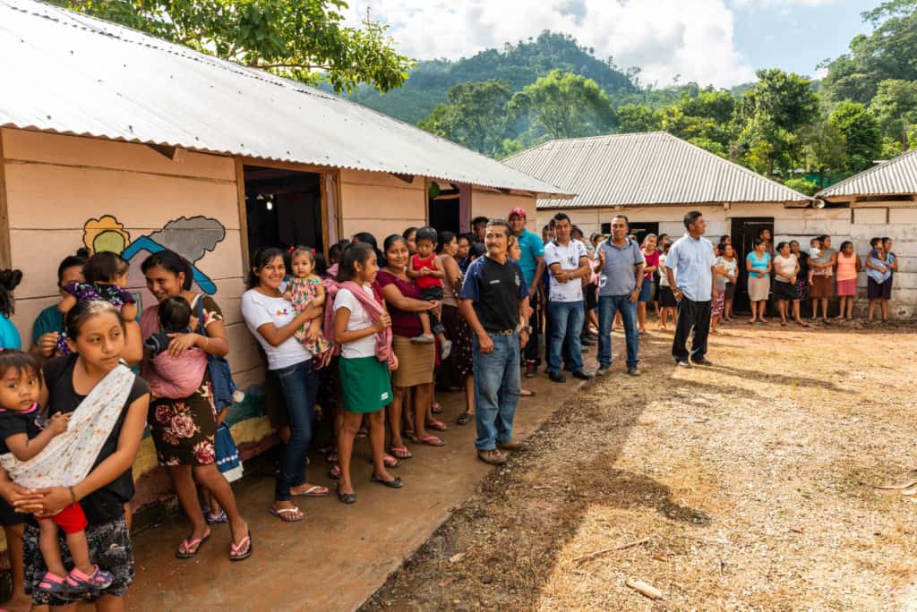 A crowd of people are standing and waiting to welcome visitors to the church. There are trees in the background.