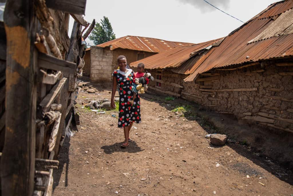 Young woman wearing a black, red, and white dress, is carrying her younger sister, wearing red, as they walk back down a dirt road to go home. There are houses all around.