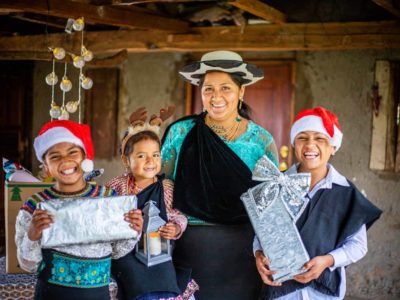 Joselin, Mayte, Jenny (mother), and Javier are inside their home. Two of the children are holding Christmas gifts and they are wearing Christmas hats.