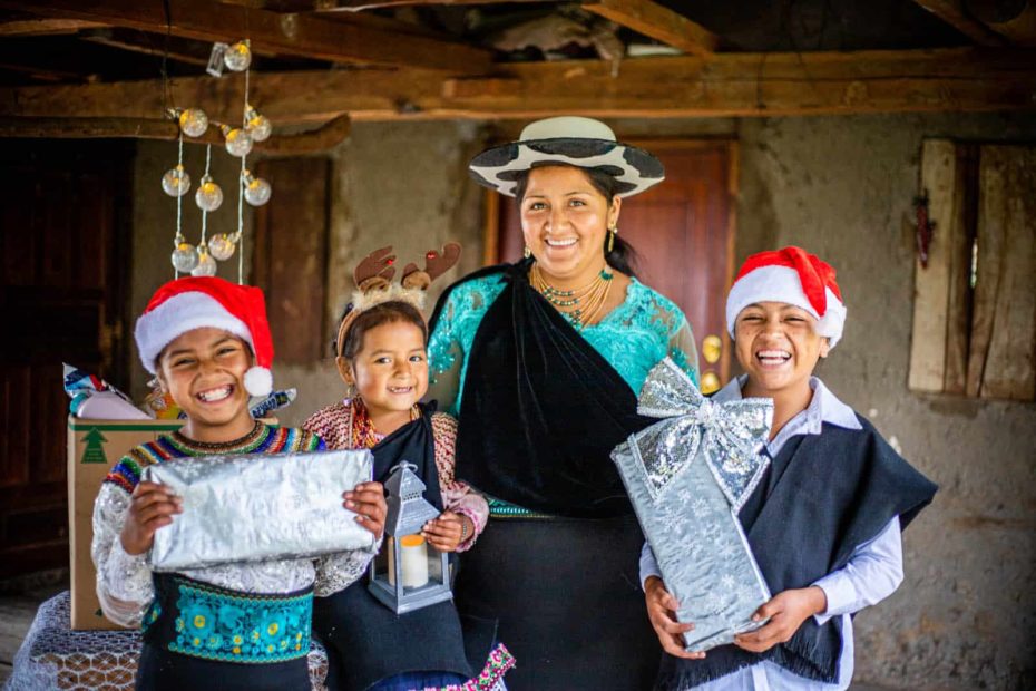 Joselin, Mayte, Jenny (mother), and Javier are inside their home. Two of the children are holding Christmas gifts and they are wearing Christmas hats.