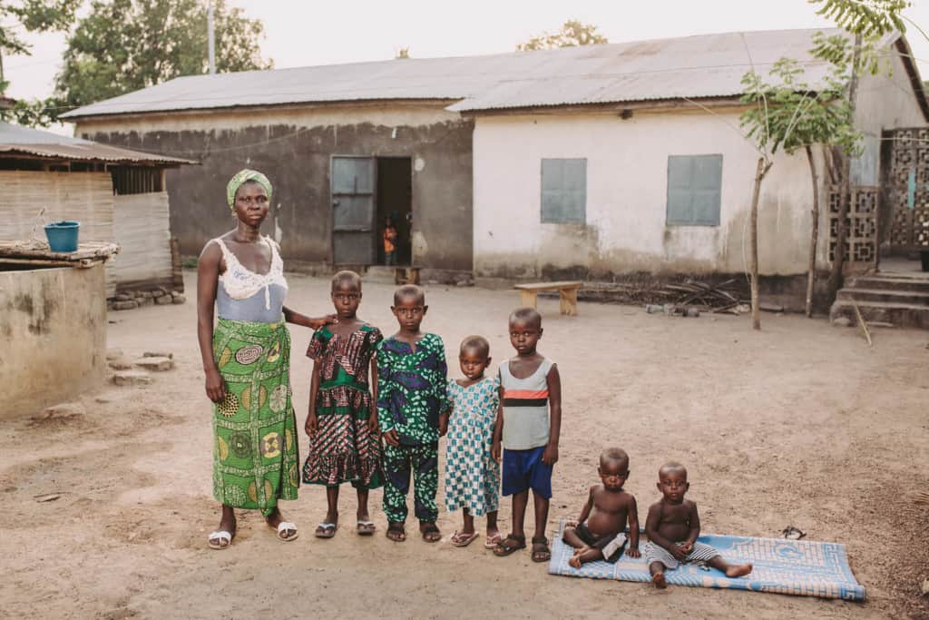 Mother wearing a white and gray sleeveless shirt a green pattern long skirt and green head scarf wrap poses outside her home near her two twin boys sitting together on a blue blanket on the ground and some of her other children standing beside her in front of their home. There is a concrete water source well in the background.