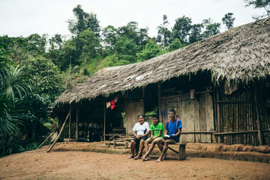 A mother, father and son sit outside in front of a wood home on a bench. They wear white, green and blue shirts. The house has a thatched roof and there are trees in the background.