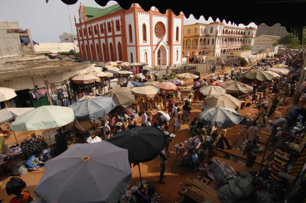 The Grand Market in Lome, Togo is in front of a large white and rust colored church. The vendors have large umbrellas over their wares and people are crowding the walkway between them.