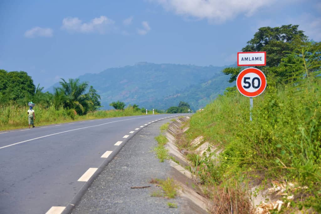 The road that leads into Etonam's town. There is a woman walking down the road and there are mountains in the background.