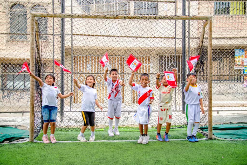 A group of children holding, waiving Peru flags. They are standing, jumping on green grass, a green soccer field. They are laughing and smiling. A soccer goal and buildings are in the background.