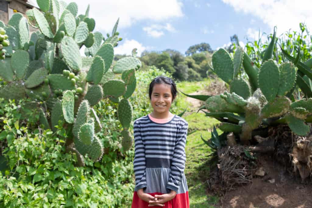 Girl standing in the middle of two cactus, demonstrating that she was victorious over the thorns of death. She is wearing a blue and black striped shirt, and smiles at the camera.